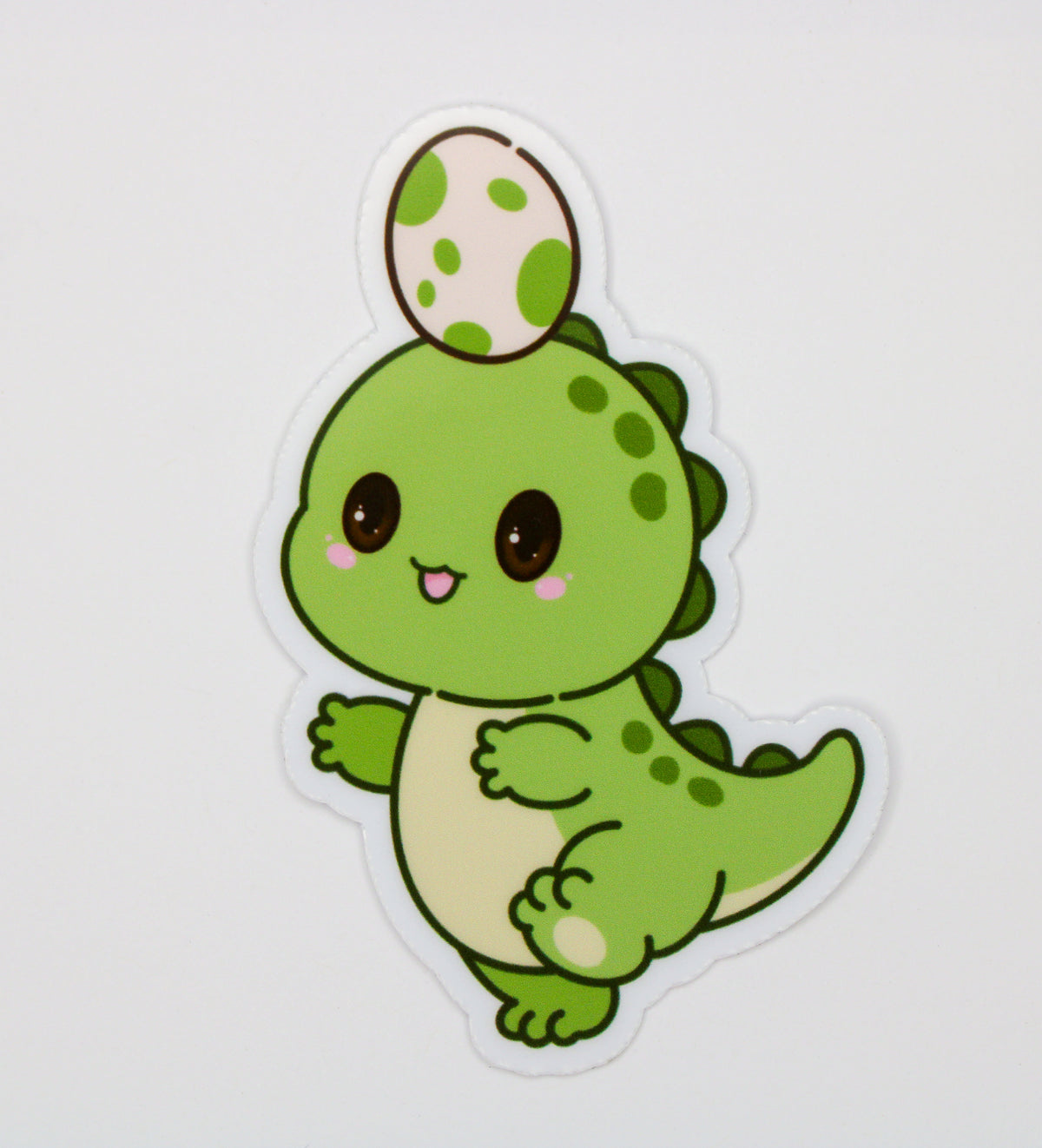 A die-cut sticker featuring a cute cartoon green dinosaur holding a speckled green and cream egg, shown on a plain light gray background.