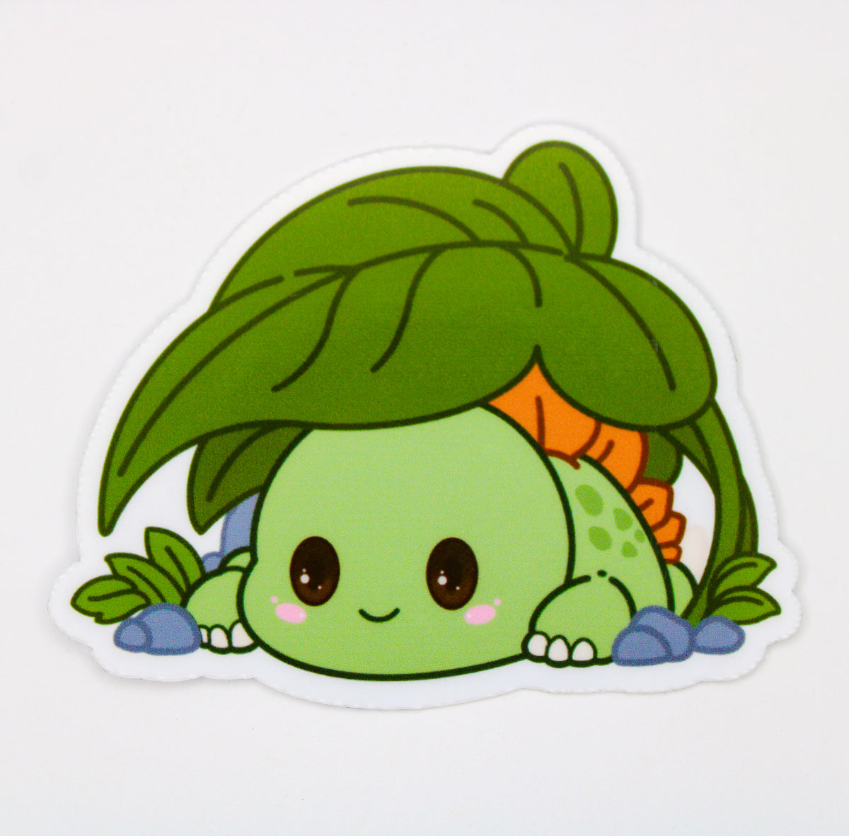 A die-cut vinyl sticker featuring a cute cartoon character resembling a green turtle or frog peeking up from behind a large green leaf, with blue stones at the base, shown on a plain white background.