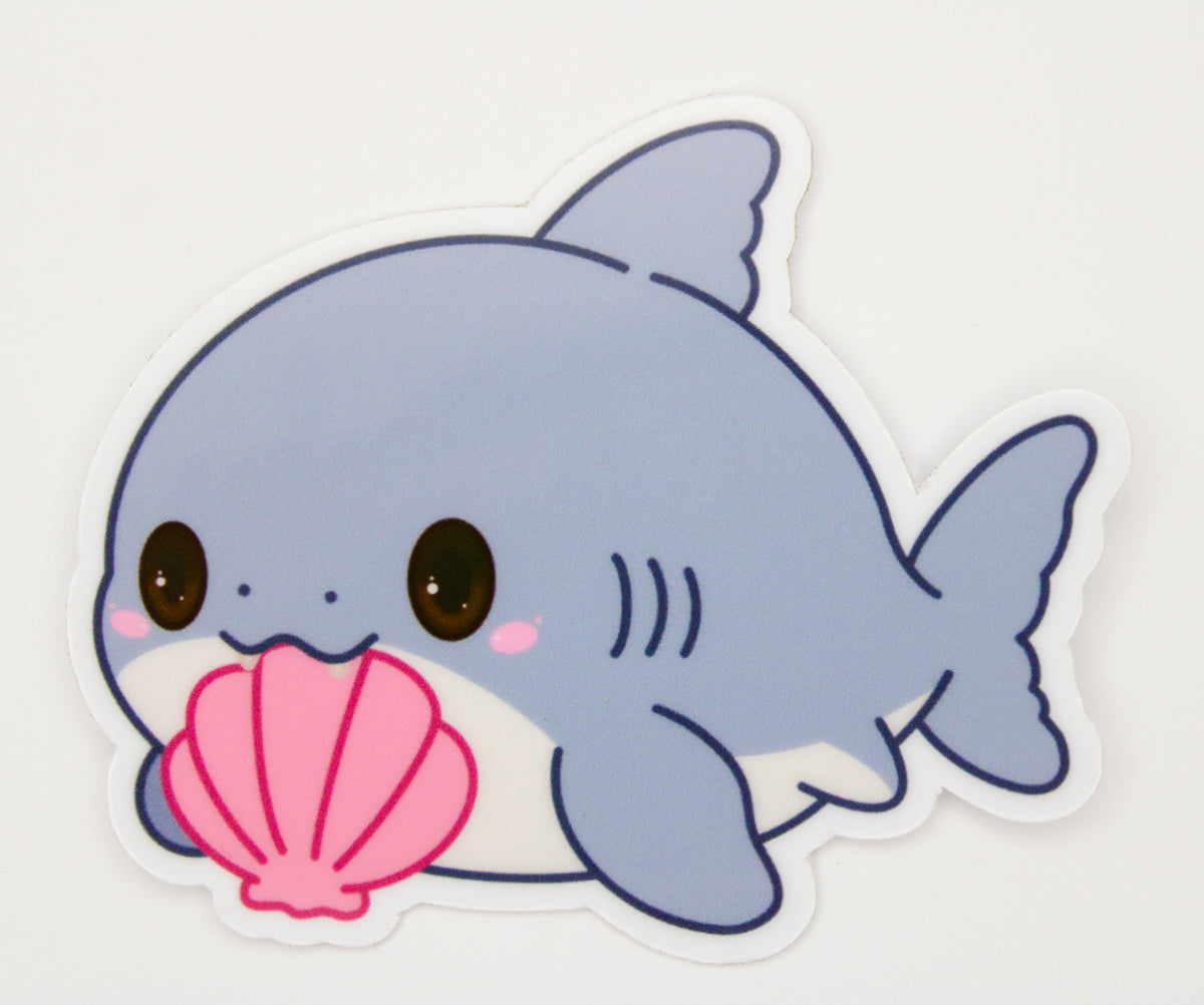A die-cut sticker featuring a cute, kawaii cartoon baby shark in light blue and white, holding a pink seashell, shown on a plain white background.