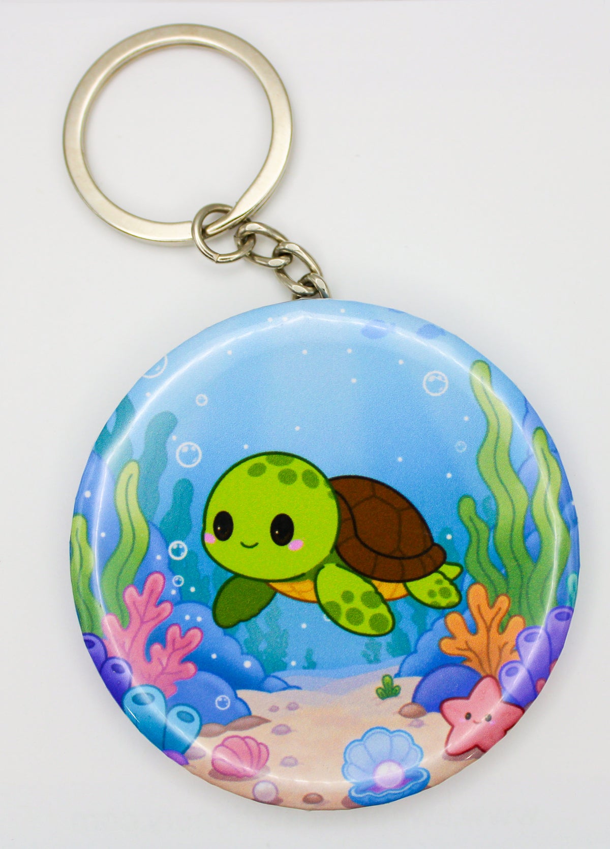 A round keychain featuring a colorful cartoon sea turtle underwater scene with coral and bubbles, attached to a gold-tone metal split ring on a white background.