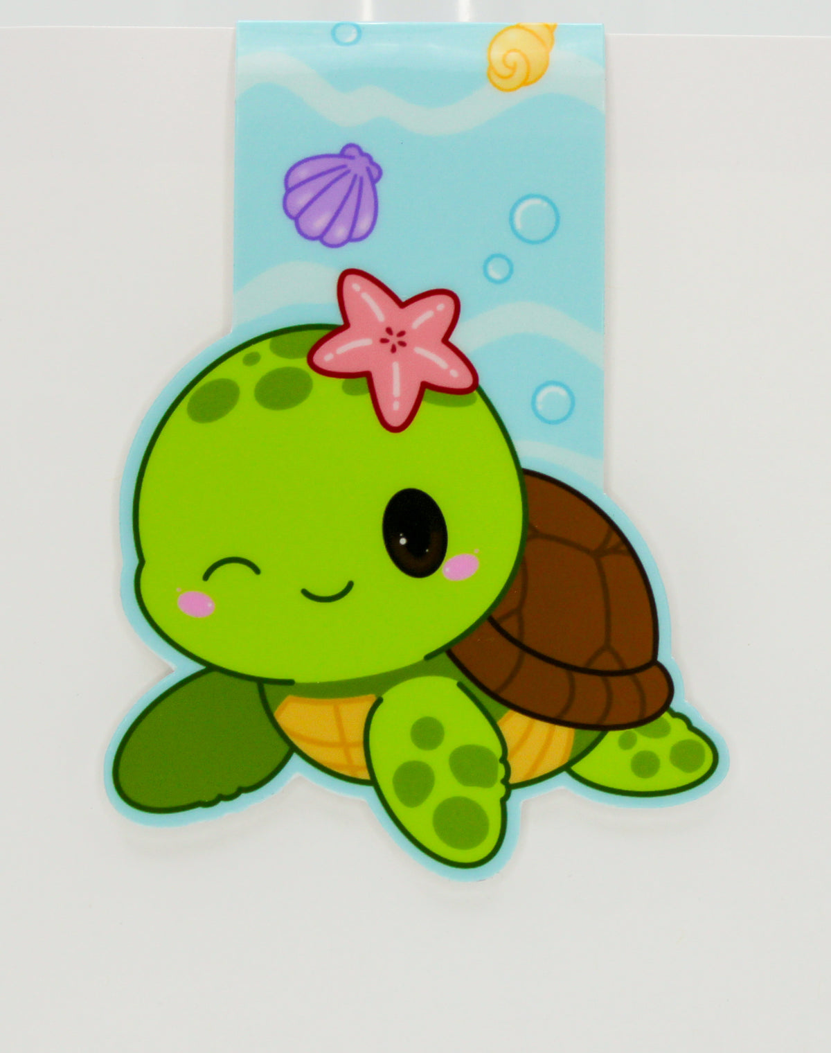 A cute cartoon sea turtle magnetic bookmark with a green body, brown shell, pink starfish on its head, and a light blue underwater-themed ribbon above it