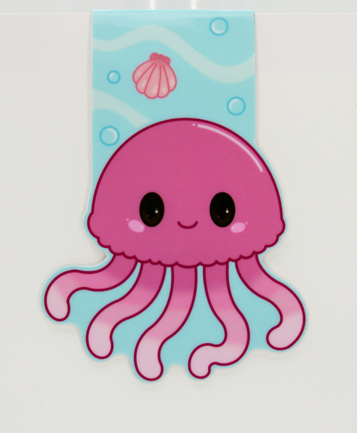 A cute pink cartoon jellyfish magnetic bookmark with black eyes and a smiling mouth, set against a light blue underwater-themed background with shells and bubbles.