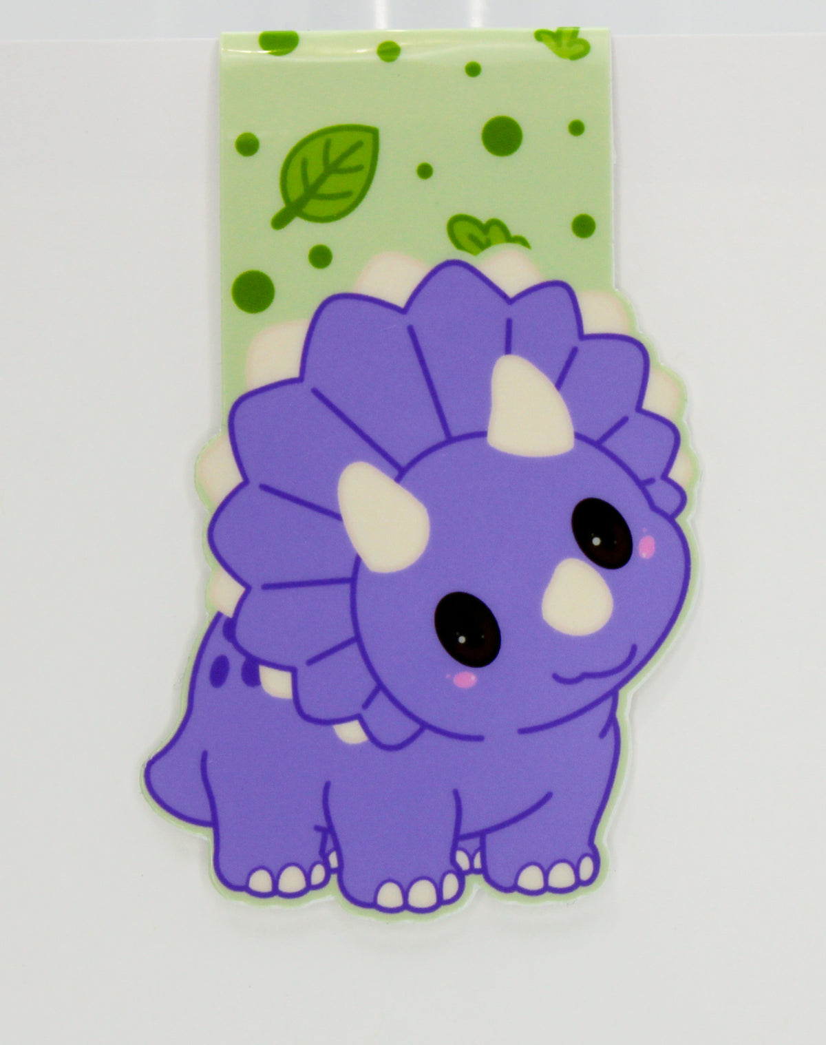 A purple cartoon Triceratops dinosaur magnetic bookmark with a light green patterned backing sheet featuring green leaves and dots.