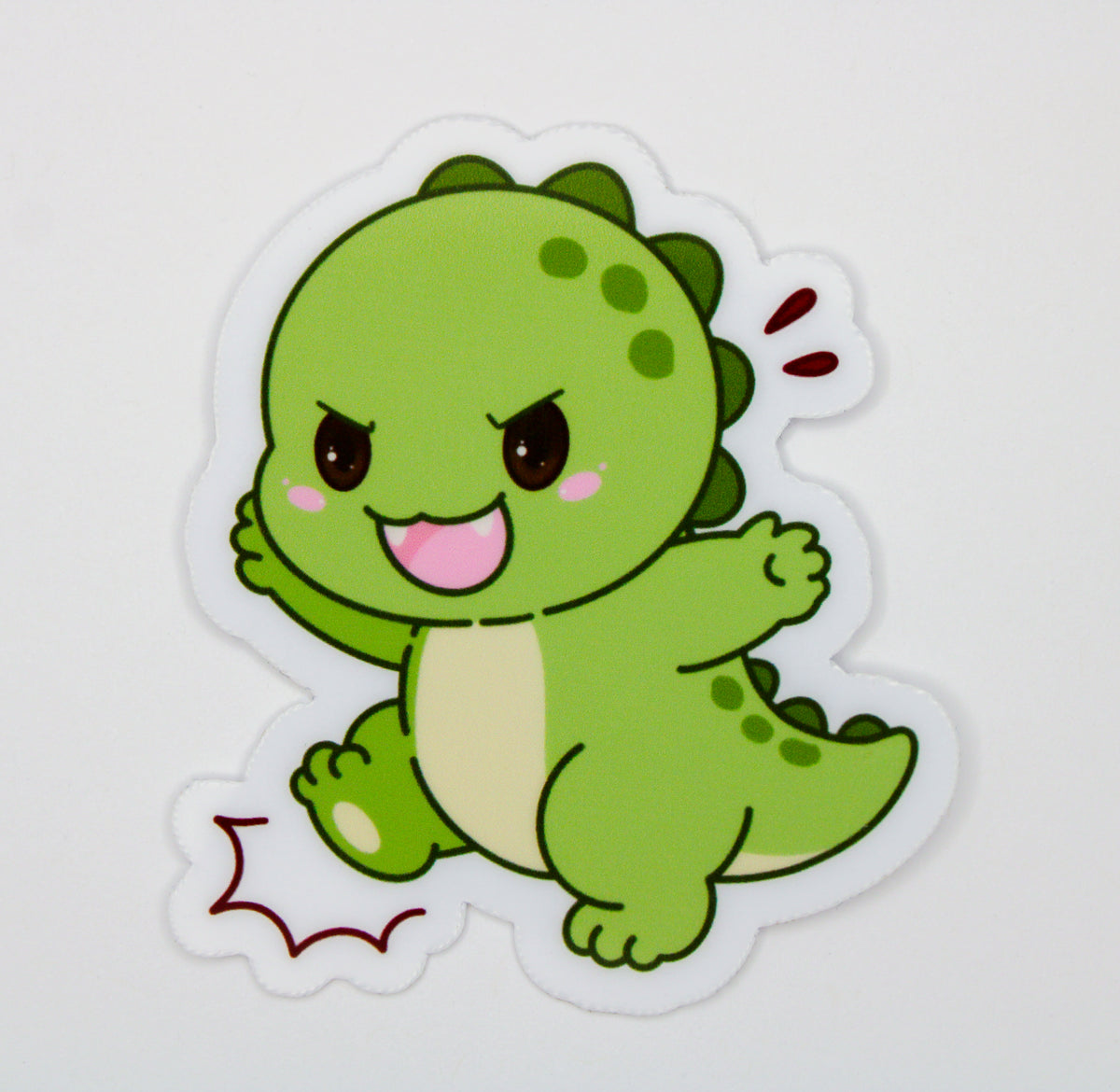 A die-cut sticker featuring a cute, cartoon-style green dinosaur character with a pink mouth and dark eyes, shown on a plain white background.