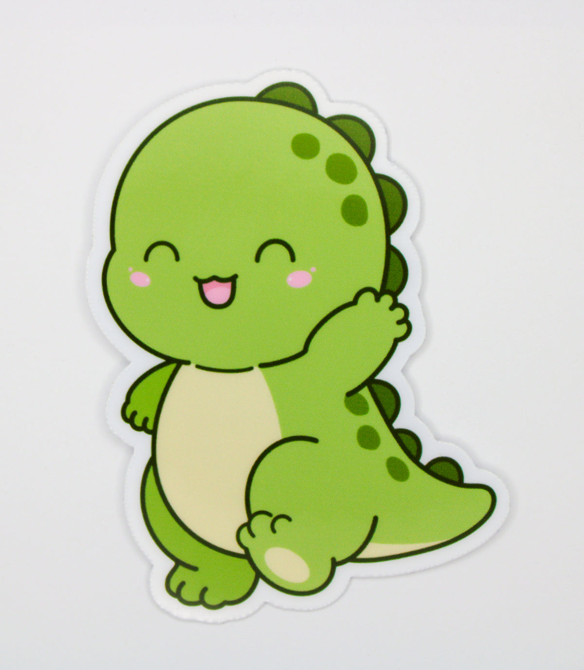 A die-cut sticker featuring a cute, smiling green cartoon dinosaur with a cream belly, shown on a plain light gray background.