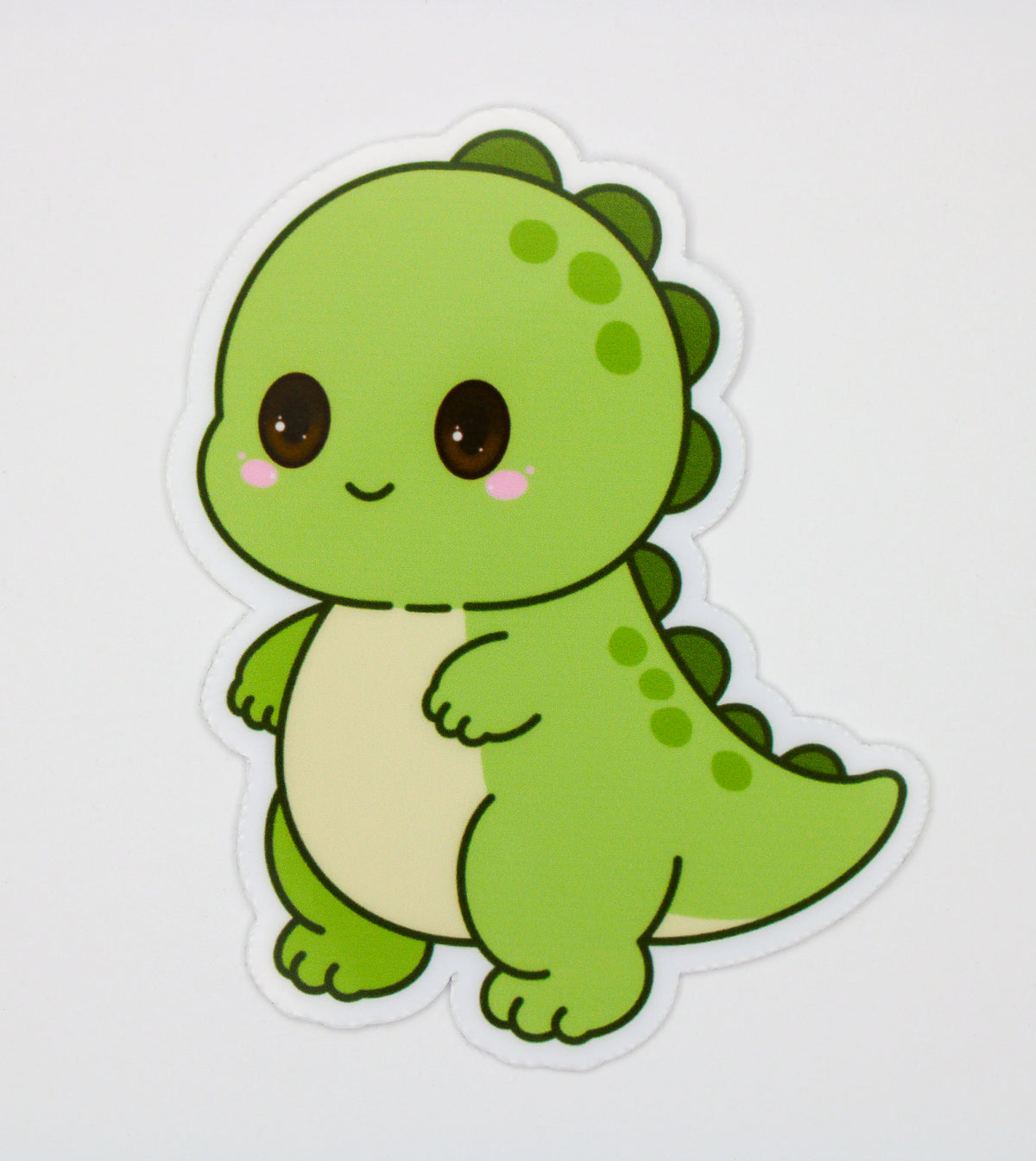 A die-cut sticker featuring a cute, cartoon-style green dinosaur with a cream-colored belly, large dark brown eyes, and pink cheeks, shown on a plain light gray background.
