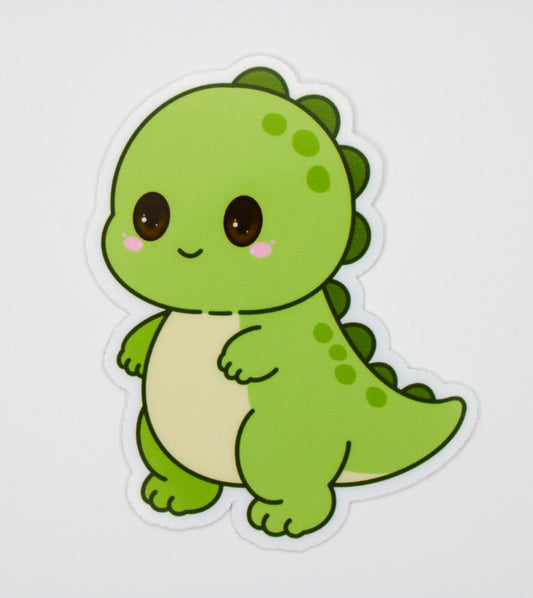 A die-cut sticker featuring a cute, cartoon-style green dinosaur with a cream-colored belly, large dark brown eyes, and pink cheeks, shown on a plain light gray background.