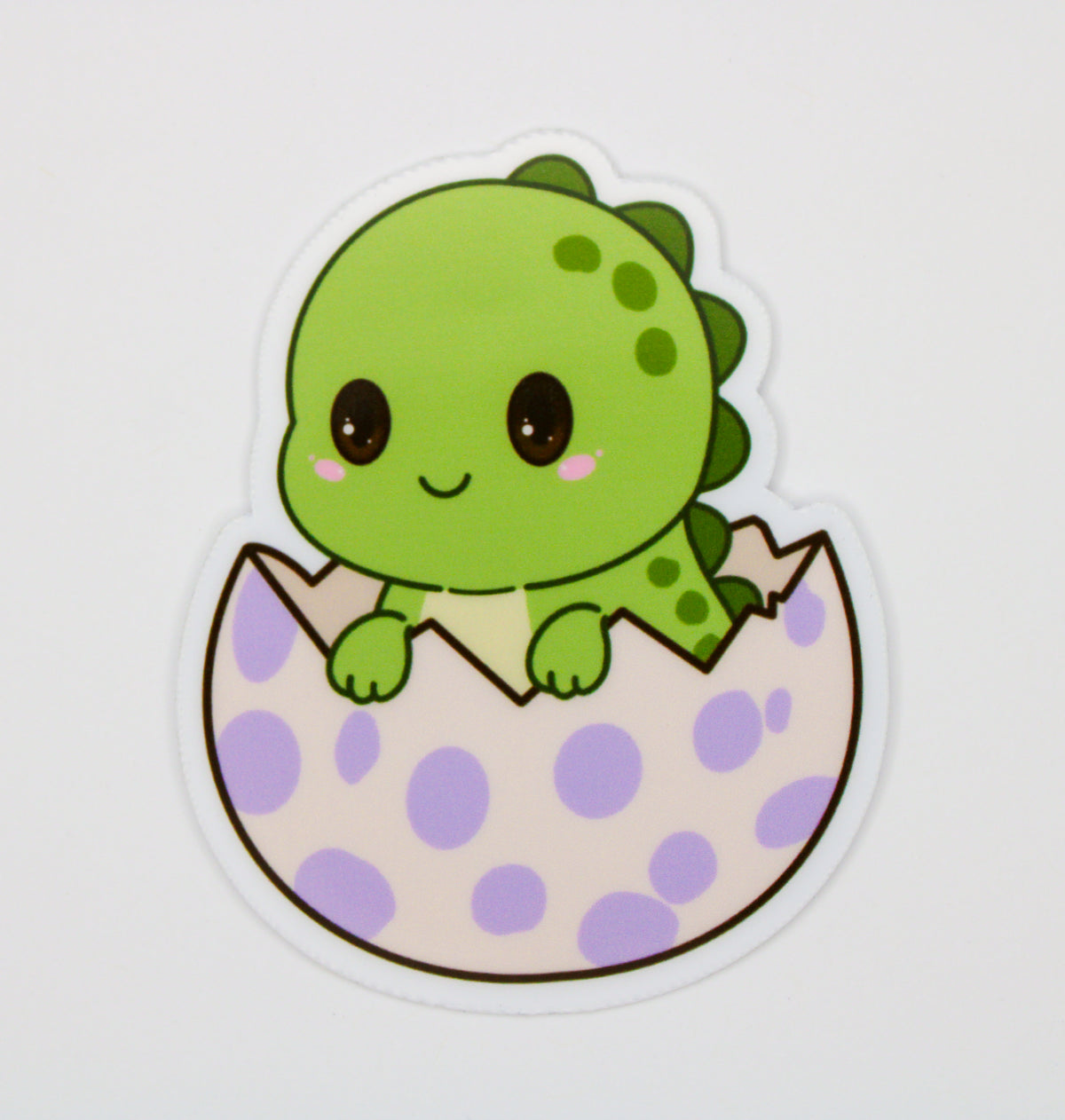 A die-cut sticker featuring a cute cartoon green dinosaur peeking out of a light purple and white polka-dotted egg, shown on a plain light gray background.