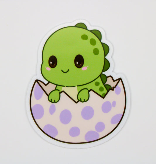 A die-cut sticker featuring a cute cartoon green dinosaur peeking out of a light purple and white polka-dotted egg, shown on a plain light gray background.