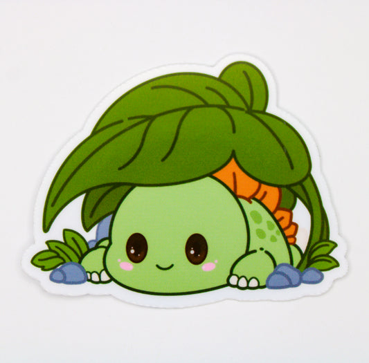 A die-cut vinyl sticker featuring a cute cartoon character resembling a green turtle or frog peeking up from behind a large green leaf, with blue stones at the base, shown on a plain white background.