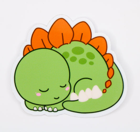 A die-cut sticker featuring a cute sleeping green dinosaur with orange spikes and white teeth, shown on a plain white background.