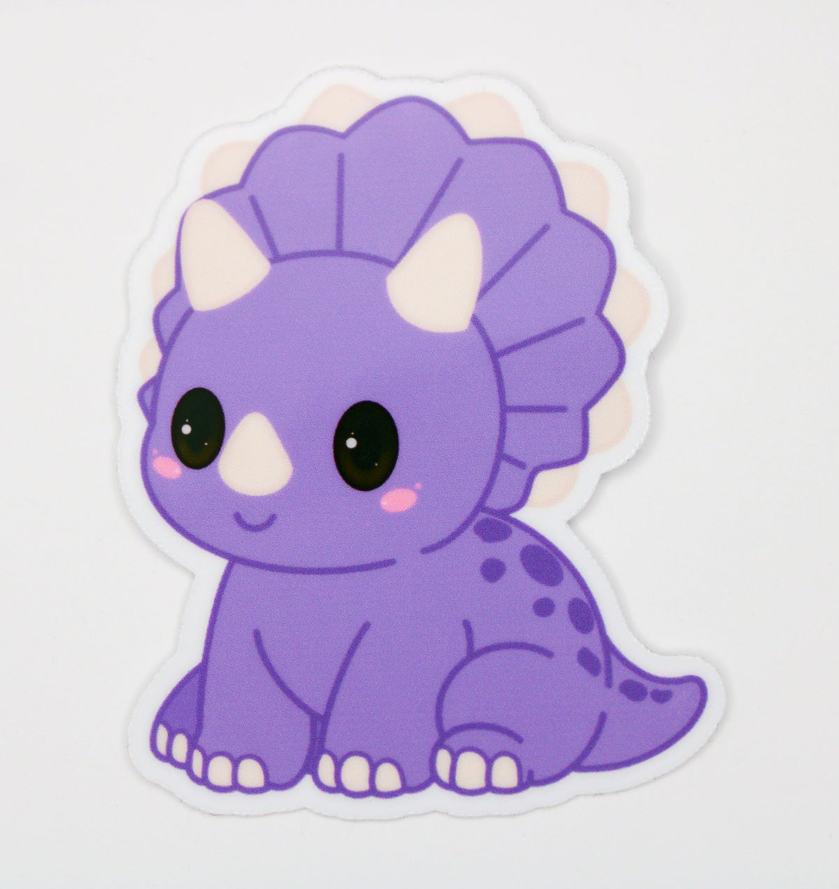 A cute die-cut sticker of a purple cartoon Triceratops dinosaur with large black eyes, pink cheeks, and cream horns, shown on a plain white background.