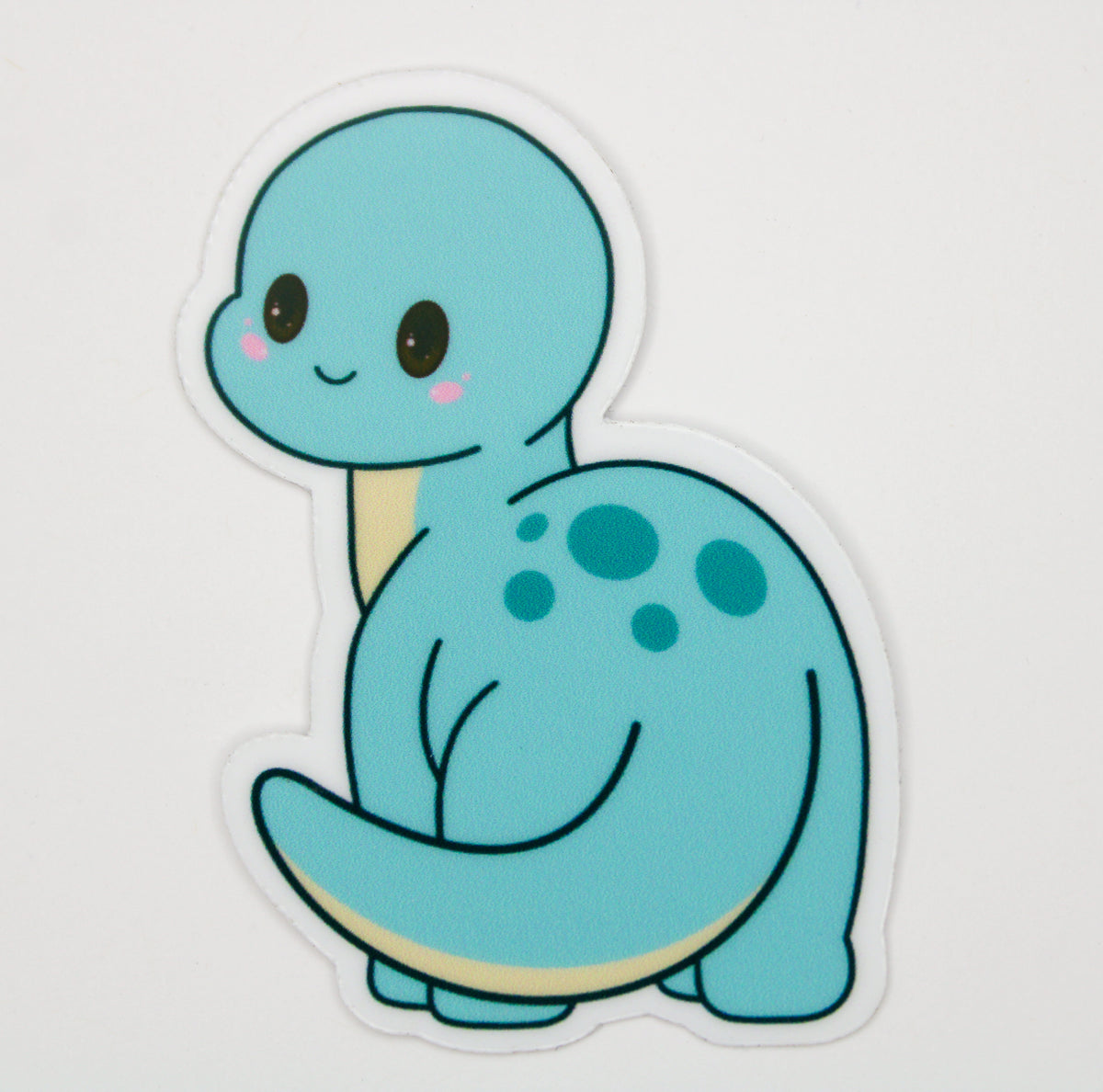 A die-cut vinyl sticker featuring a cute, cartoon-style light blue dinosaur with dark blue spots, a cream underbelly, and pink cheeks, shown on a plain white background.