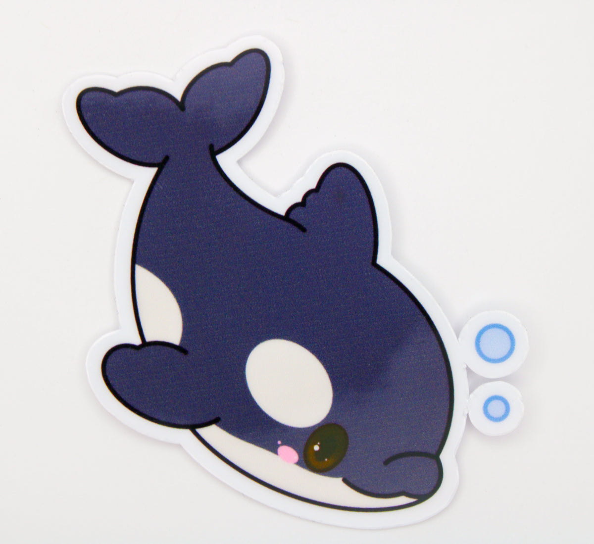A cute cartoon orca sticker on a plain white background. The sticker features a dark navy blue and white whale with a pink nose, a brown eye, and two small blue bubbles on the right side.