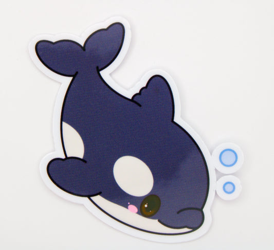 A cute cartoon orca sticker on a plain white background. The sticker features a dark navy blue and white whale with a pink nose, a brown eye, and two small blue bubbles on the right side.