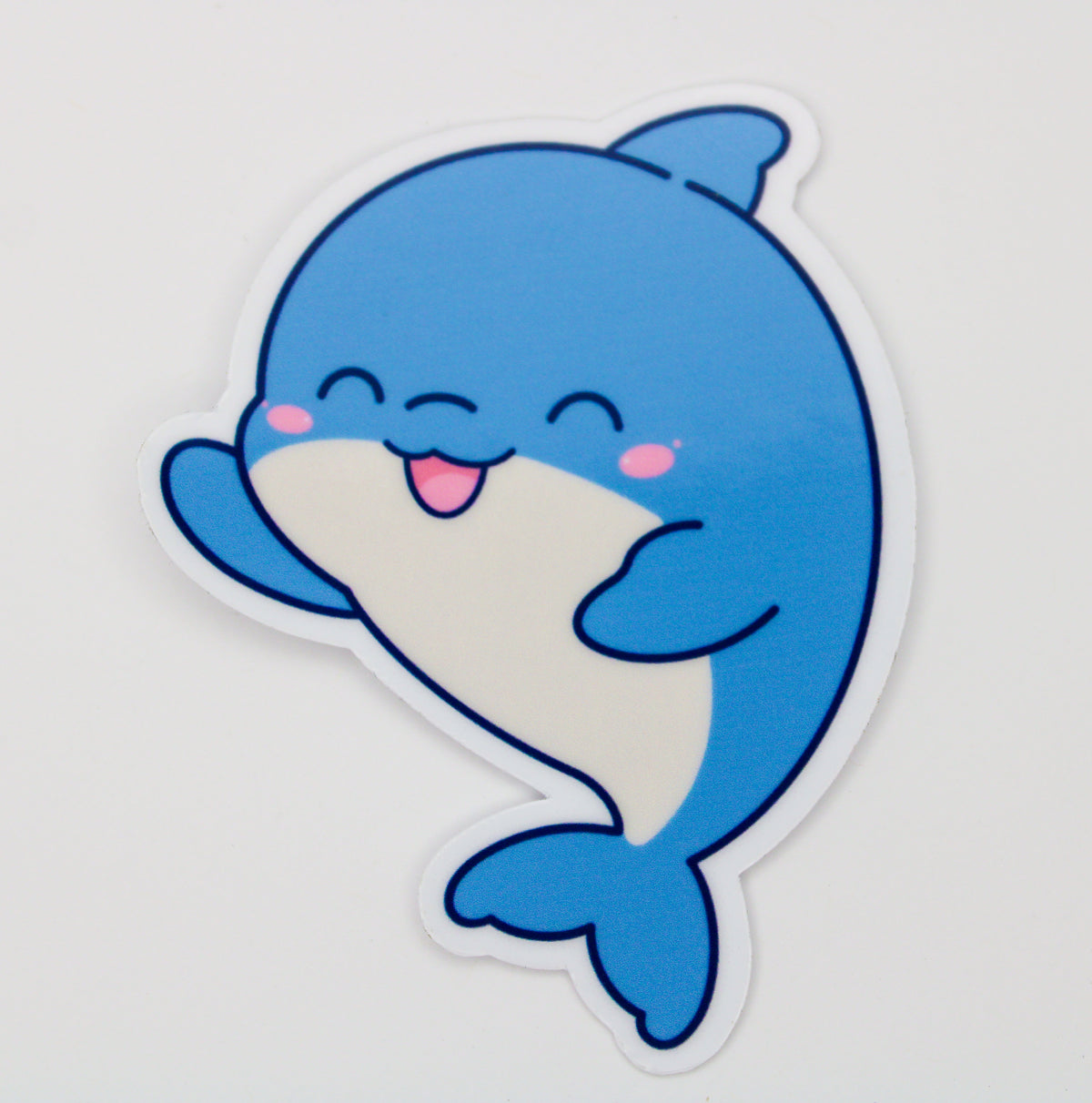 A die-cut sticker featuring a cute, smiling cartoon blue dolphin with a white belly, pink cheeks, and a pink tongue, shown on a plain light background.