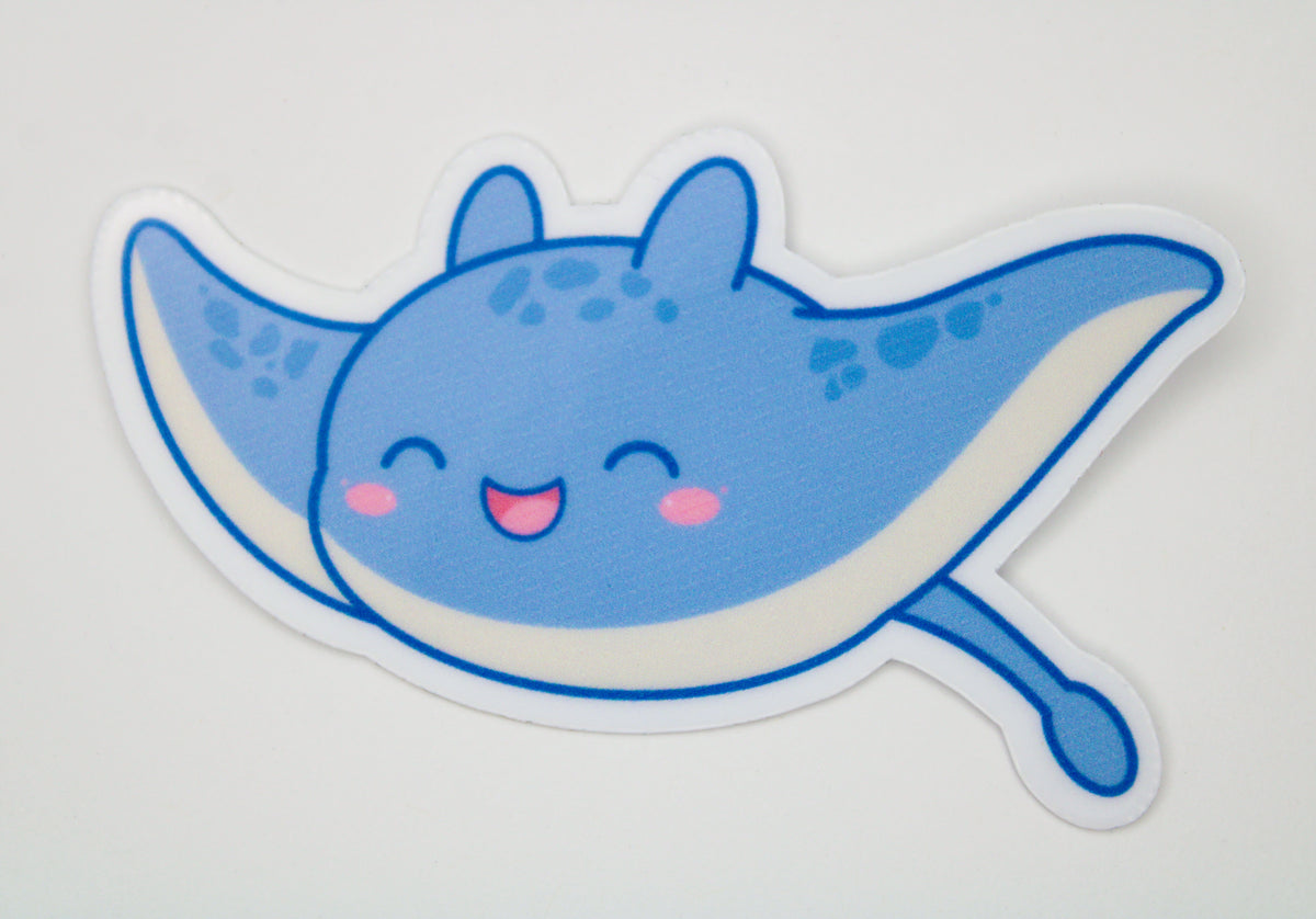 A die-cut vinyl sticker of a cute blue cartoon manta ray with pink cheeks and a smiling mouth, shown on a plain light gray background.