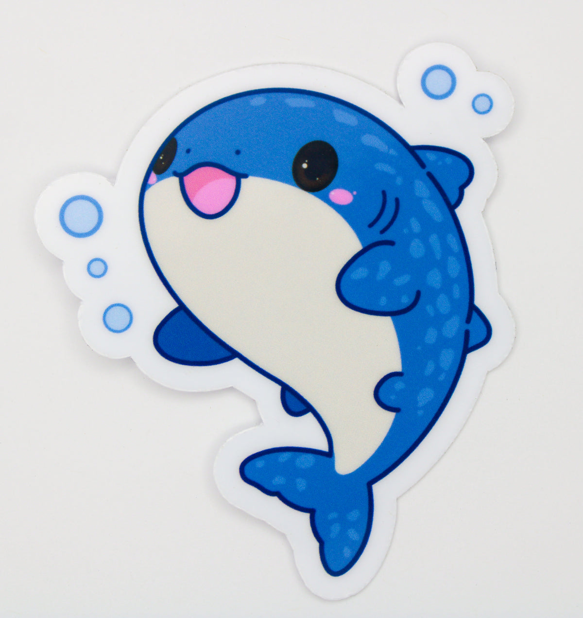 A die-cut vinyl sticker featuring a cute cartoon blue shark with a white belly, pink tongue, and blue bubbles on a light gray background.
