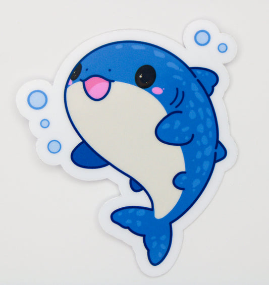 A die-cut vinyl sticker featuring a cute cartoon blue shark with a white belly, pink tongue, and blue bubbles on a light gray background.
