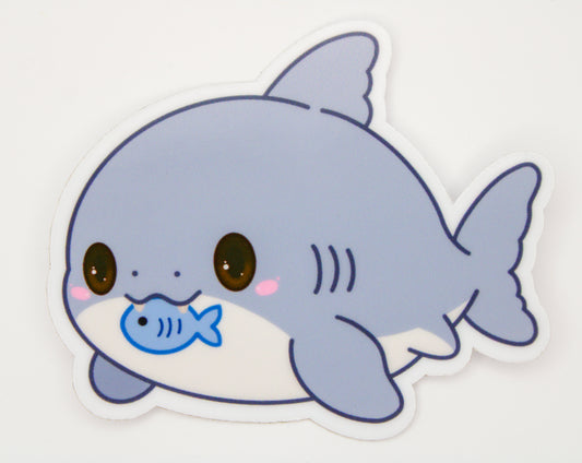 A die-cut sticker featuring a cute cartoon shark with a blue fish in its mouth, shown on a plain white background.