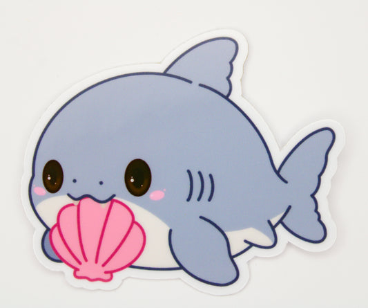 A die-cut sticker featuring a cute, kawaii cartoon baby shark in light blue and white, holding a pink seashell, shown on a plain white background.