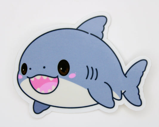 A die-cut vinyl sticker of a cute cartoon baby shark, featuring a light blue-gray body, white underbelly, pink mouth, and large brown eyes, shown on a plain white background.