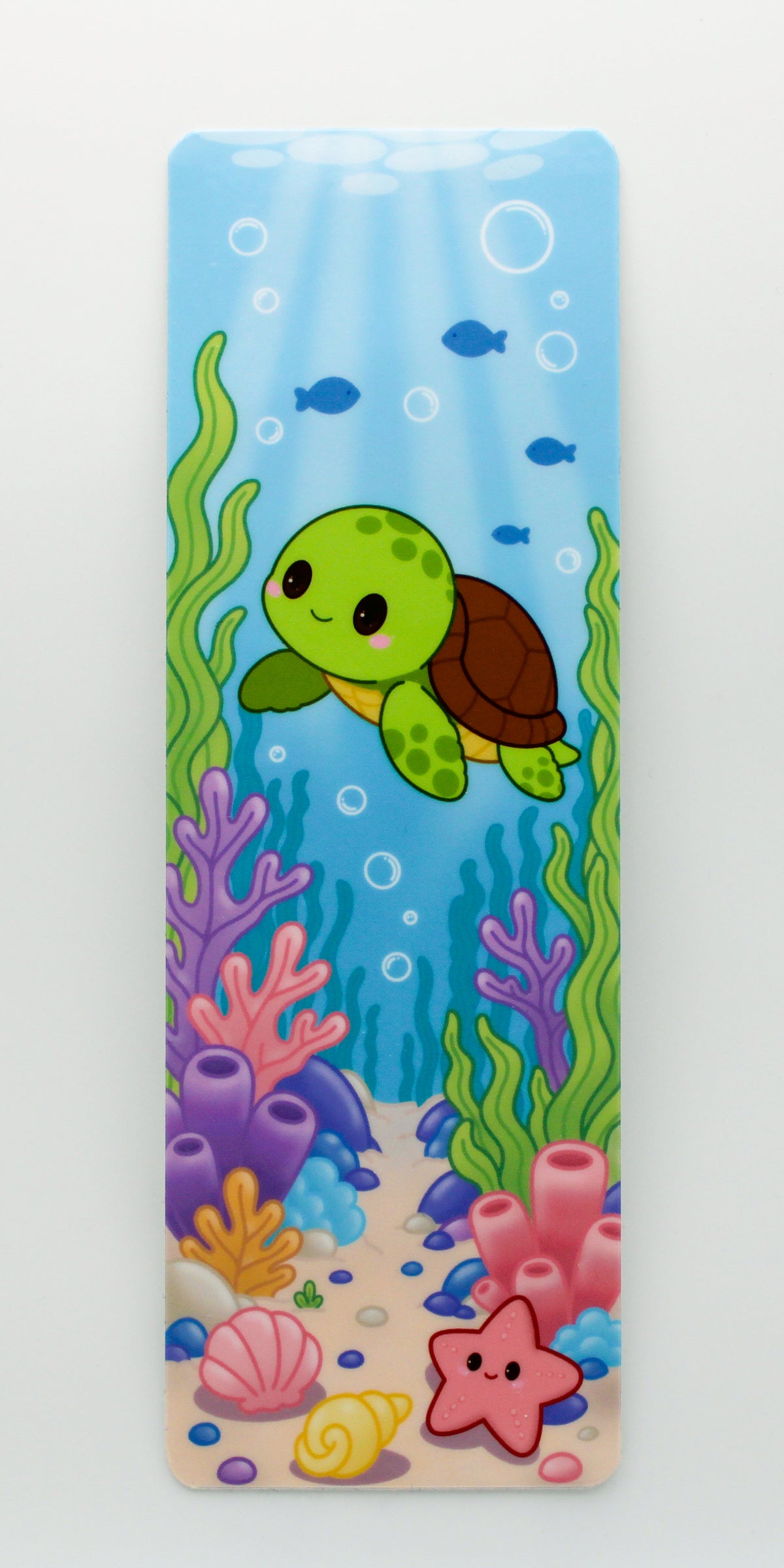 A vertical rectangular bookmark featuring a colorful cartoon underwater scene with a green sea turtle swimming among coral, seaweed, bubbles, and a sandy seabed.