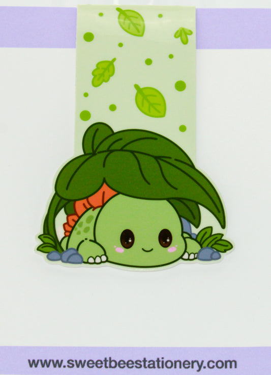 A cute cartoon magnetic bookmark of a green baby dinosaur-like character peeking under a large green leaf, with a pale green leaf-patterned strip above and a white background.
