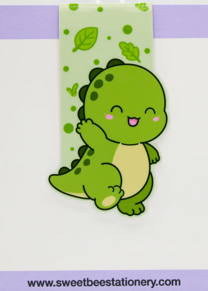A cute cartoon dinosaur hair clip by Sweet Bee Stationery, featuring a green smiling dinosaur with a cream belly and pink cheeks, attached to a light green patterned backing card.