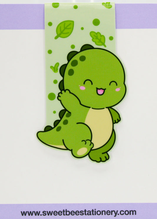 A cute cartoon dinosaur hair clip by Sweet Bee Stationery, featuring a green smiling dinosaur with a cream belly and pink cheeks, attached to a light green patterned backing card.