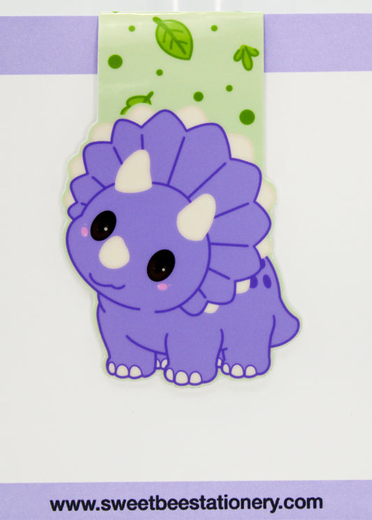 A cute magnetic bookmark of a purple cartoon triceratops dinosaur, shown on a light purple and white background with a green patterned backing card