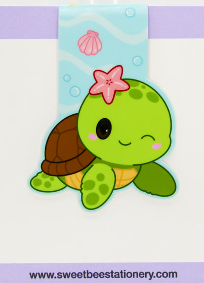 A cute cartoon sea turtle magnetic bookmark with a green body, brown shell, pink starfish on its head, and a light blue underwater-themed ribbon above it