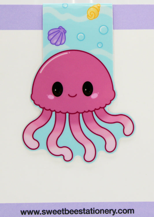 A cute pink cartoon jellyfish magnetic bookmark with black eyes and a smiling mouth, set against a light blue underwater-themed background with shells and bubbles. 