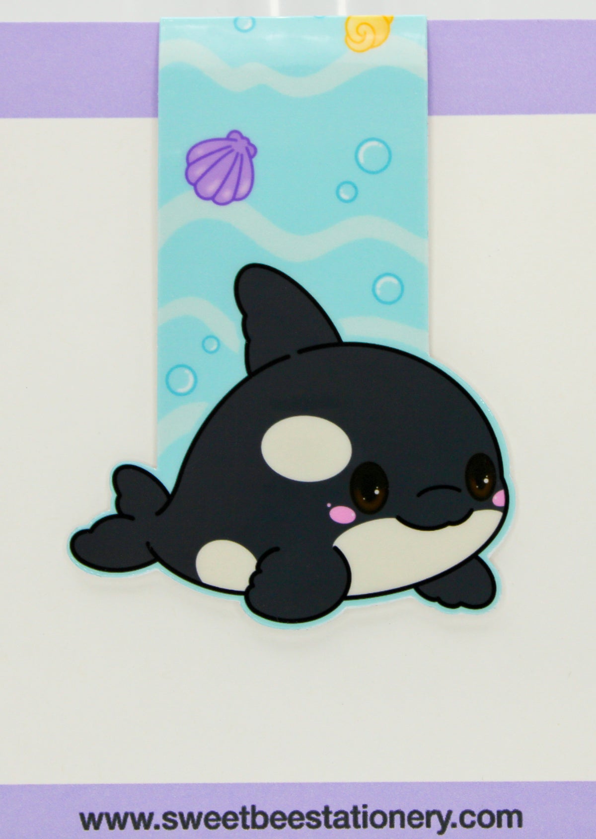 A cute chibi-style orca magnetic bookmark shown on a white background with a purple strip at the top and a website watermark at the bottom. The orca is black and white with a small pink accent, positioned in front of a vertical underwater-themed blue panel with wavy lines, bubbles, a shell, and a sun.