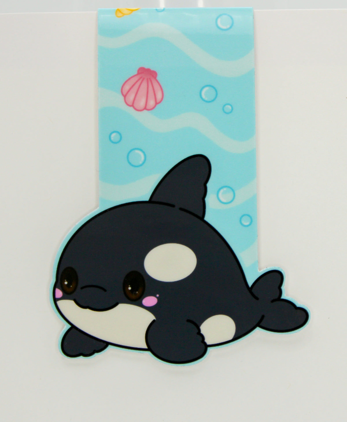A cute chibi-style orca magnetic bookmark shown on a white background with a purple strip at the top and a website watermark at the bottom. The orca is black and white with a small pink accent, positioned in front of a vertical underwater-themed blue panel with wavy lines, bubbles, a shell, and a sun.