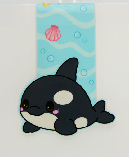 A cute chibi-style orca magnetic bookmark shown on a white background with a purple strip at the top and a website watermark at the bottom. The orca is black and white with a small pink accent, positioned in front of a vertical underwater-themed blue panel with wavy lines, bubbles, a shell, and a sun.