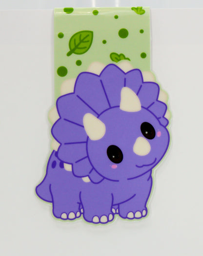 A purple cartoon Triceratops dinosaur magnetic bookmark with a light green patterned backing sheet featuring green leaves and dots.
