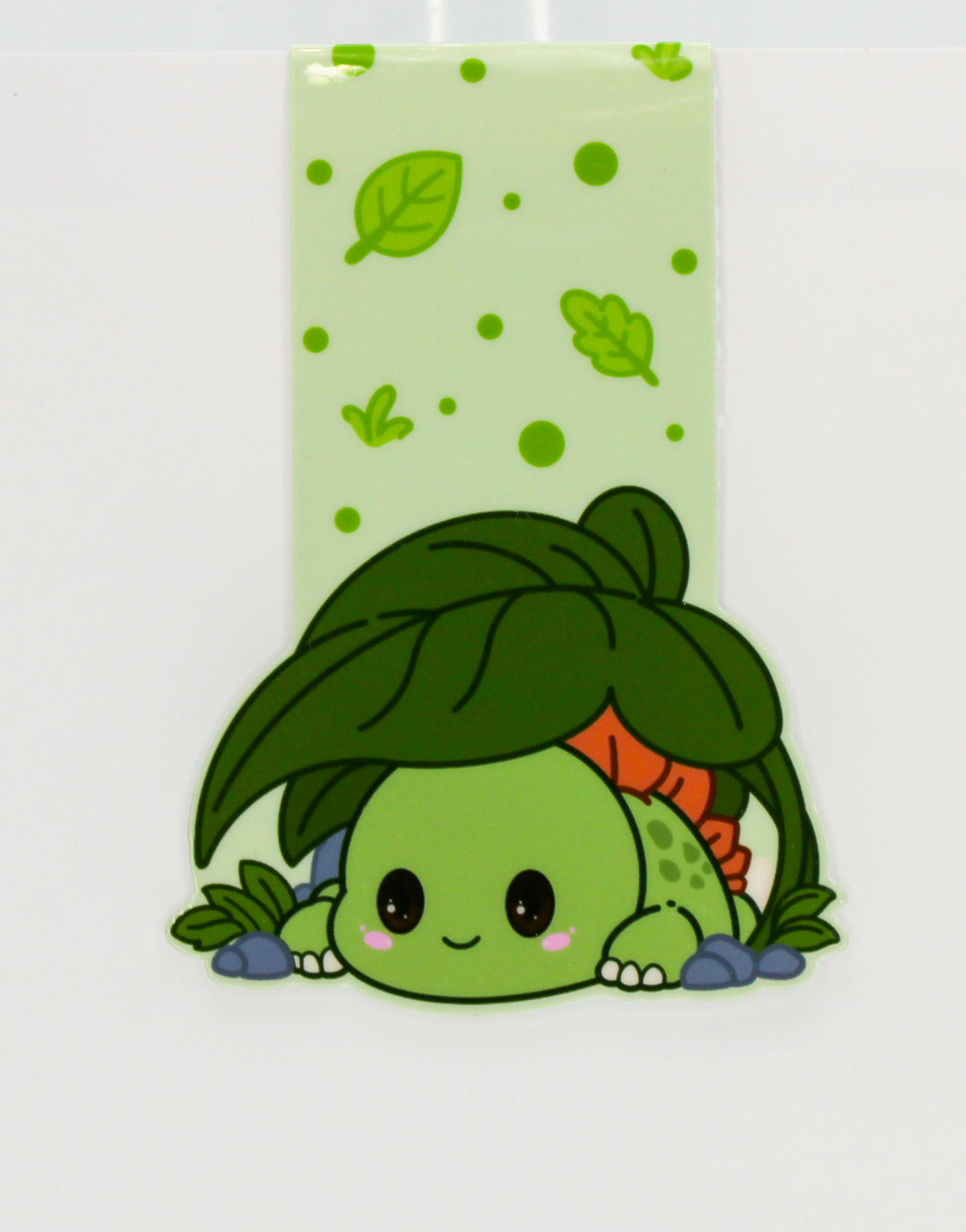 A cute cartoon magnetic bookmark of a green baby dinosaur-like character peeking under a large green leaf, with a pale green leaf-patterned strip above and a white background.